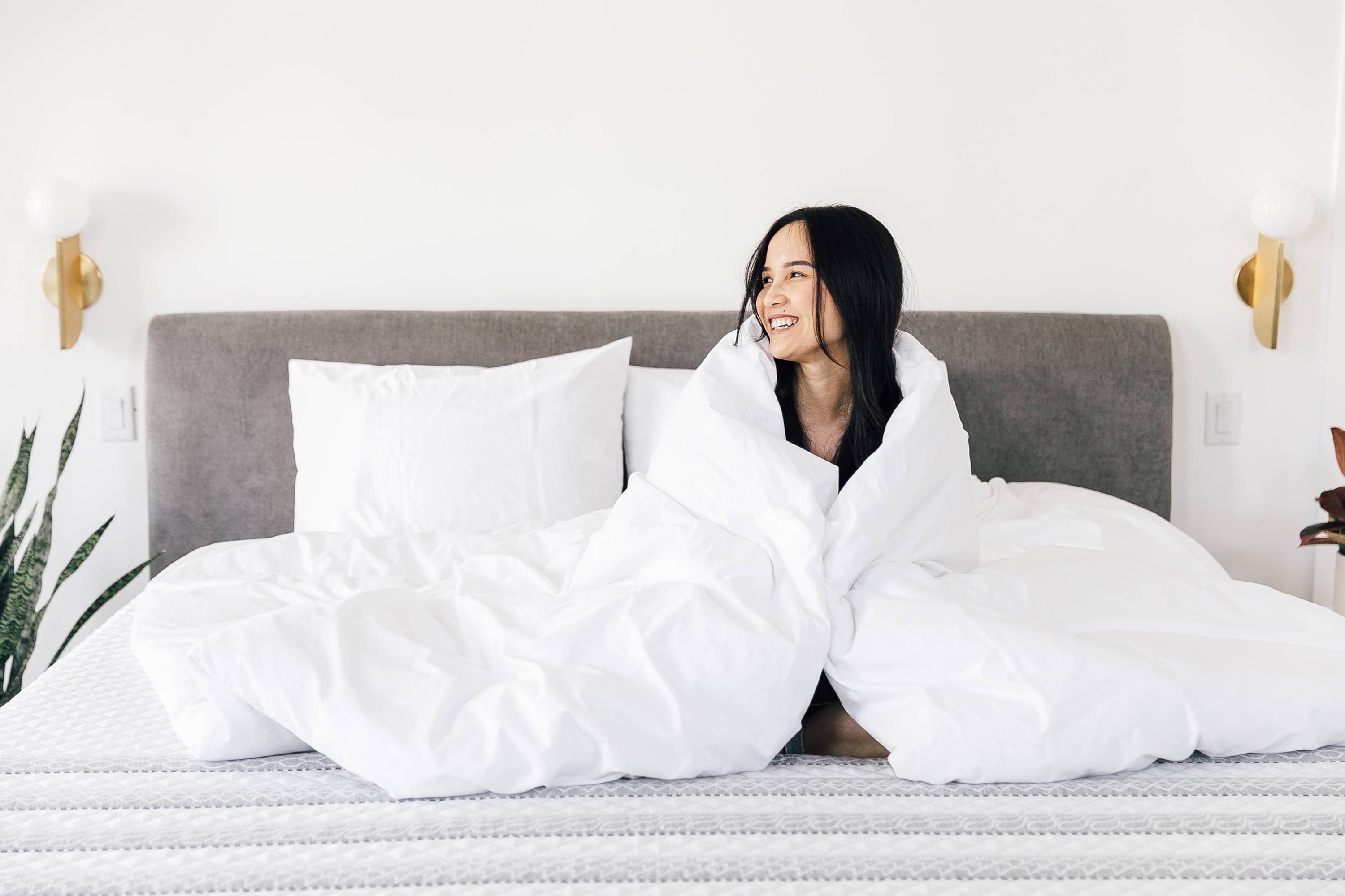 A lady smiling on a Leesa mattress wrapped in a white comforter.